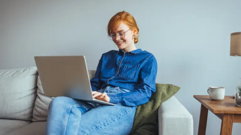 A woman working on her laptop from the comfort of her couch with a smile on her face