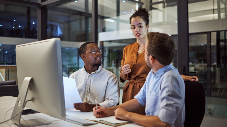 A woman standing over two men while they work in front of a desktop computer within an office setting