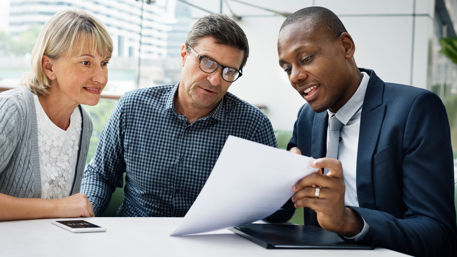 A life insurance agent in a black suit sharing some documents with two clients of his, a man and a woman.