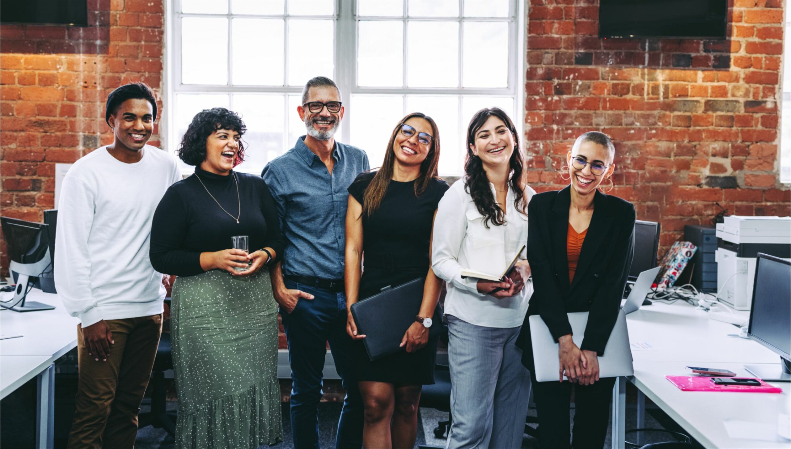 A group of six co-workers smiling and laughing in a row while waiting for a picture to be taken