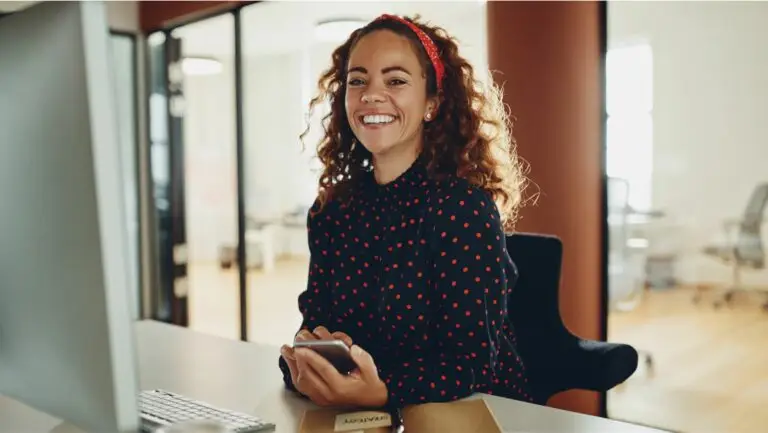 A smiling woman holding a phone, sitting at a desk in front of a laptop