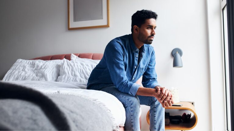 A man sitting on the edge of the bed in work clothes, looking defeated