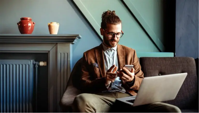 A man sitting on a couch with a laptop on his lap and a phone in his hand.