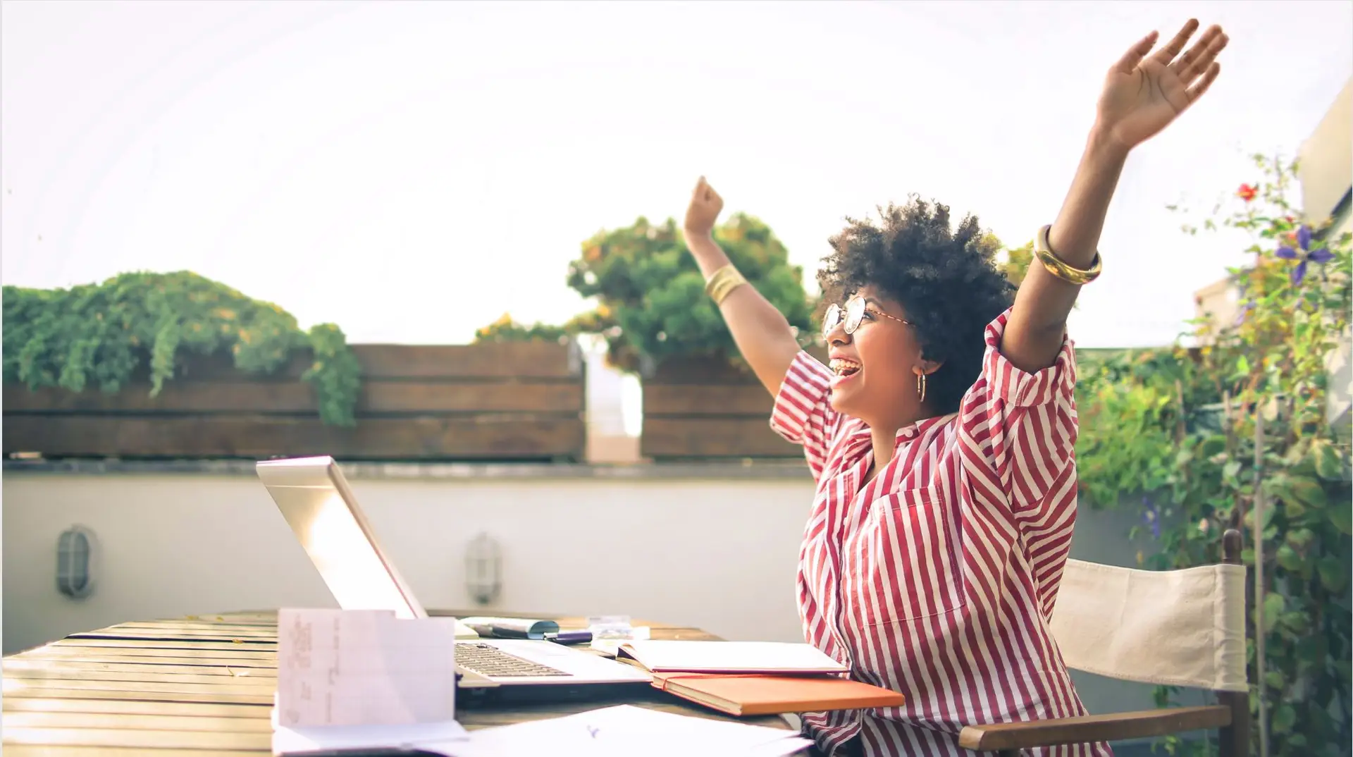 A woman working outside on her laptop throwing her arms into the air in celebration