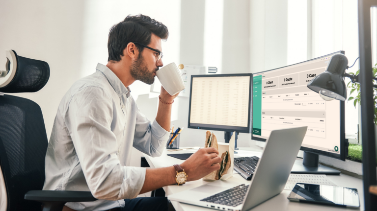 Man working from a desk with a sandwich in one hand and a cup of coffee in the other while staring at a screen with Quility Navigator on it.