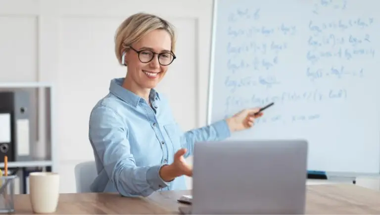 A woman meeting with someone virtually through her laptop while pointing to a whiteboard with written text behind her