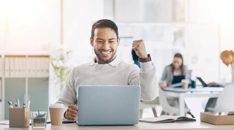 A man celebrating a personal win in front of a laptop on his desk in an office setting