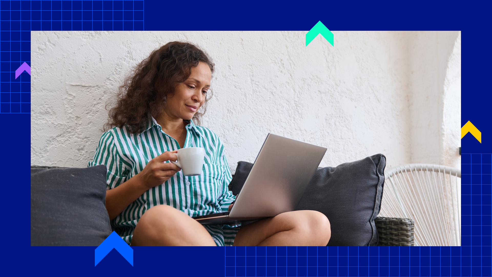 A woman on a couch, wearing pajamas, and working from a laptop with a coffee mug in hand.