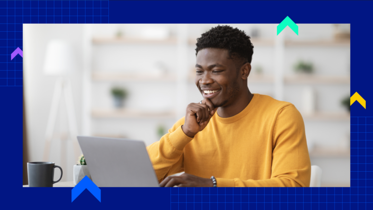 A man is smiling in a yellow sweatshirt in front of his laptop