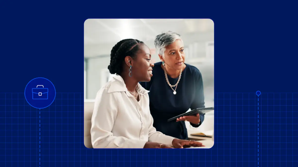 Two women in a professional setting, one holding a tablet and speaking while the other listens attentively, both smiling.